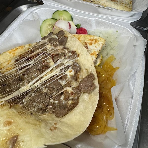 Cheesy steak quesadilla with stringy melted cheese, caramelized onions, and fresh vegetables in a to-go container.