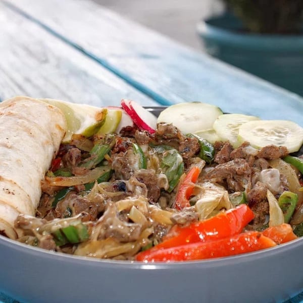 A bowl of Mexican fajita-style beef with peppers, onions, and a tortilla on a blue wooden table.