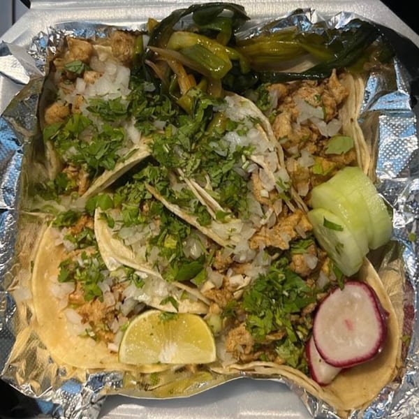 Four chicken tacos with cilantro, onions, lime, cucumber, and radish in a foil-lined container.
