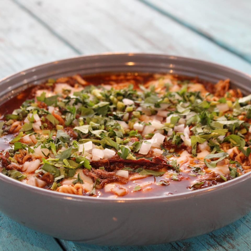 Close-up of a bowl of birria ramen, garnished with cilantro and diced onions, on a blue wooden table.